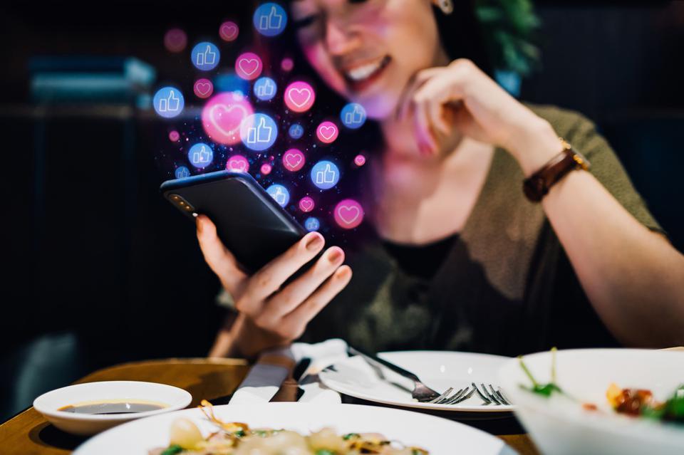 Smiling young Asian woman using smartphone on social media network application while having meal in the restaurant, viewing or giving likes, love, comment, friends and pages. Social media addiction concept